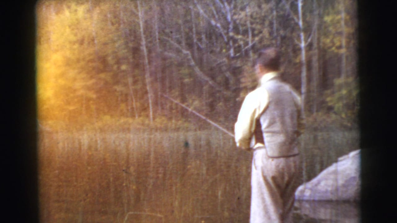 1956 8mm frame from Miami metadata showing a man fishing at a lake edge, with reeds and trees behind him and warm exposure flare at left.