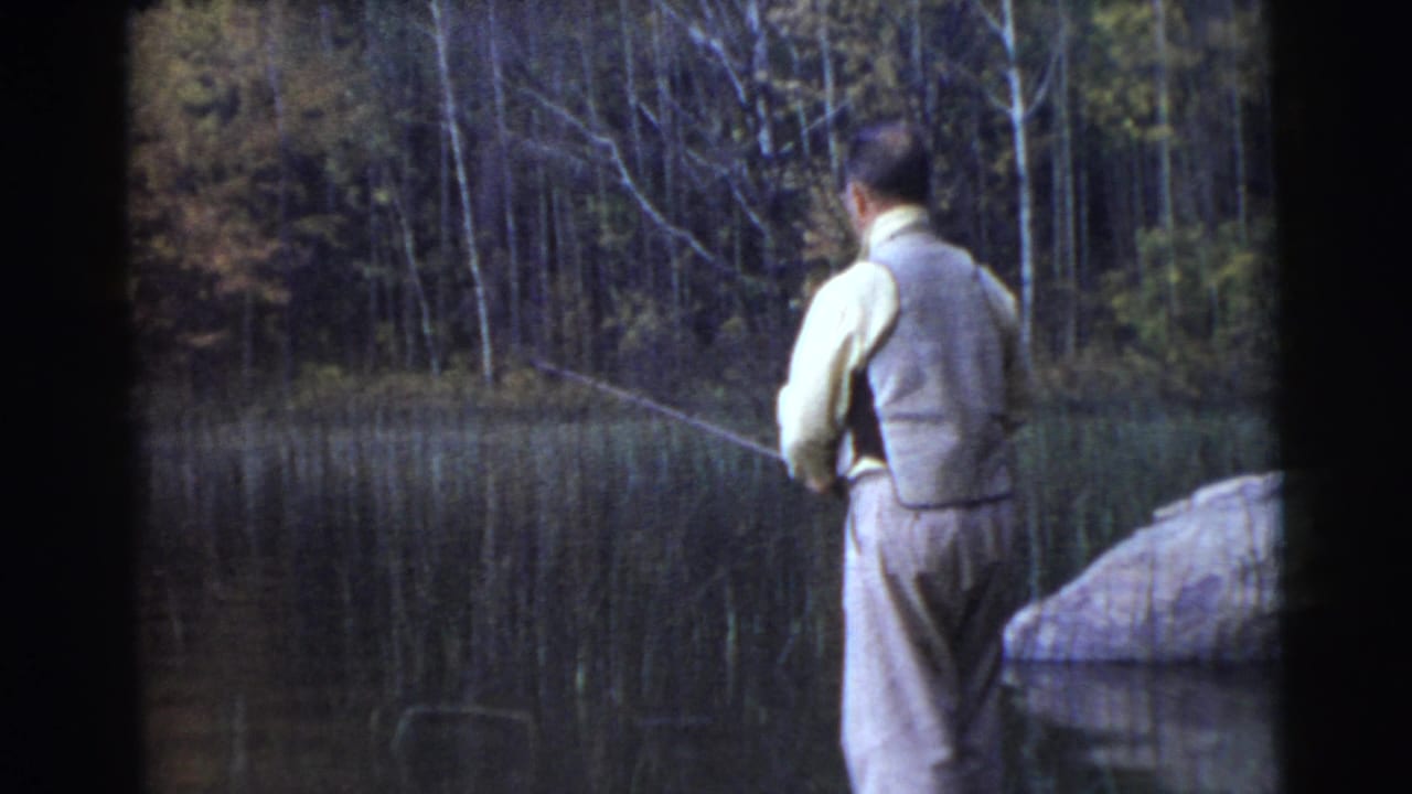 1956 8mm frame from Miami metadata showing a man fishing from the lakeside, framed by calm water, reeds, rocks, and autumn-toned trees.