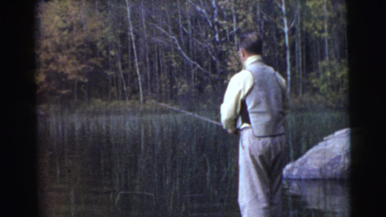 Man seen from behind fishing beside calm water with reeds and trees beyond.