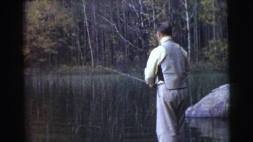 1956 8mm home-movie frame of a man fishing from a wooded waterside edge, with reeds and autumn-toned trees in view.