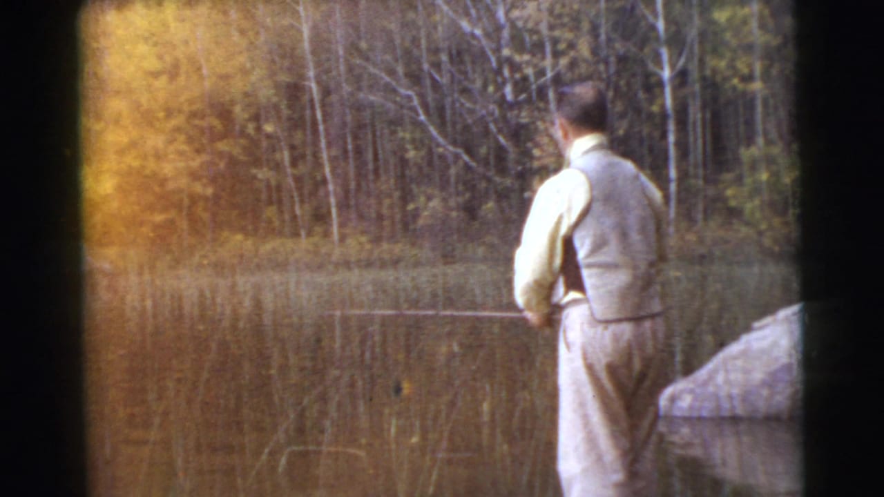 Man in light clothing holds a fishing rod near still water and trees.
