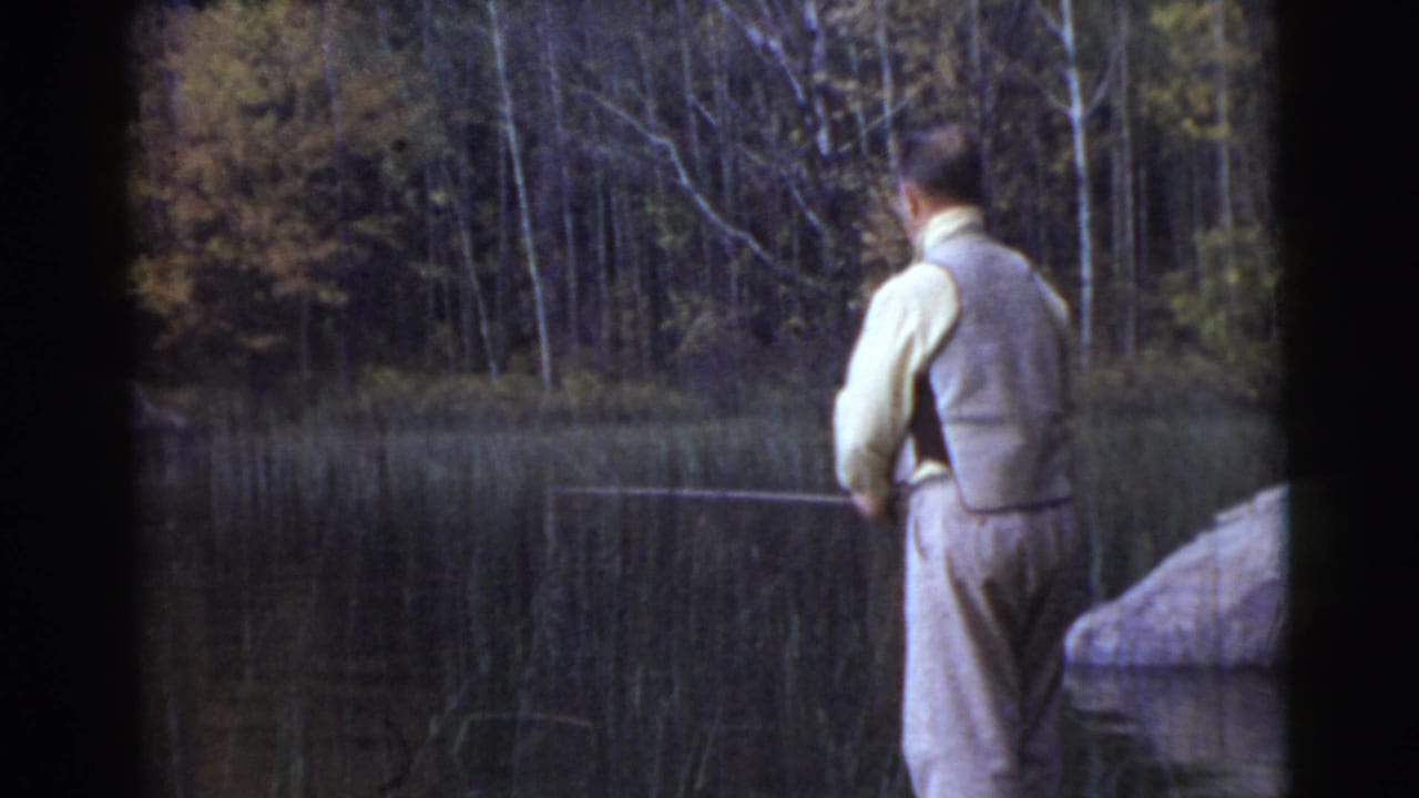 Vintage 8mm frame of the same man fishing from the bank, with a dark wooded background and calm water in front of him.