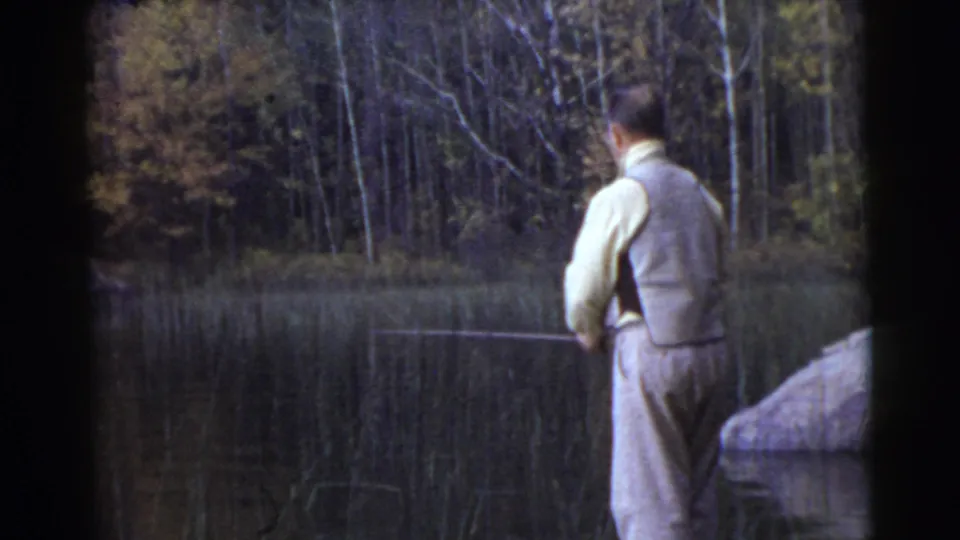 Vintage 8mm frame of the same man fishing from the bank, with a dark wooded background and calm water in front of him.