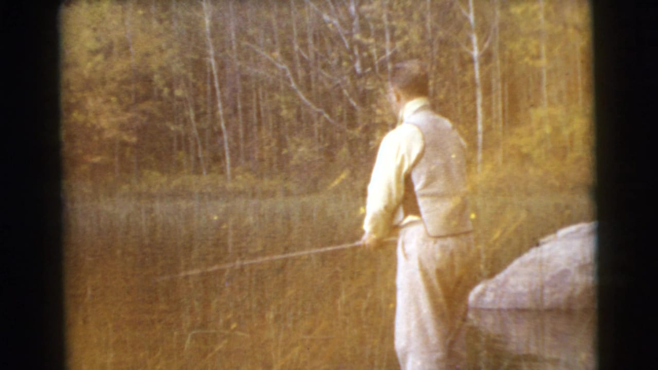 1956 home-movie frame of a man fishing at the edge of calm water, with warm exposure, reeds, and trees behind the lake.