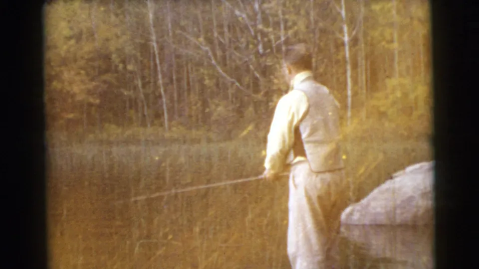 1956 home-movie frame of a man fishing at the edge of calm water, with warm exposure, reeds, and trees behind the lake.