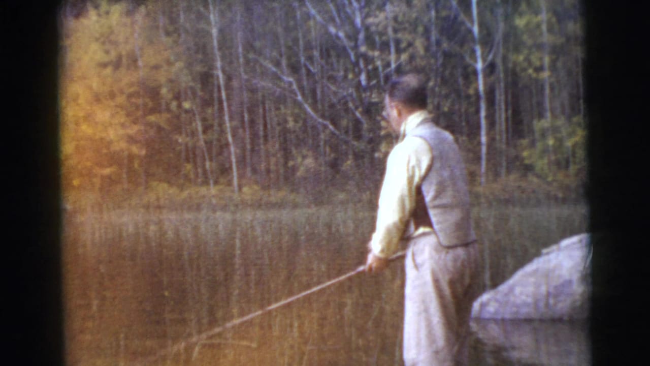 Man in light clothing holds a fishing rod at the edge of a calm lake.