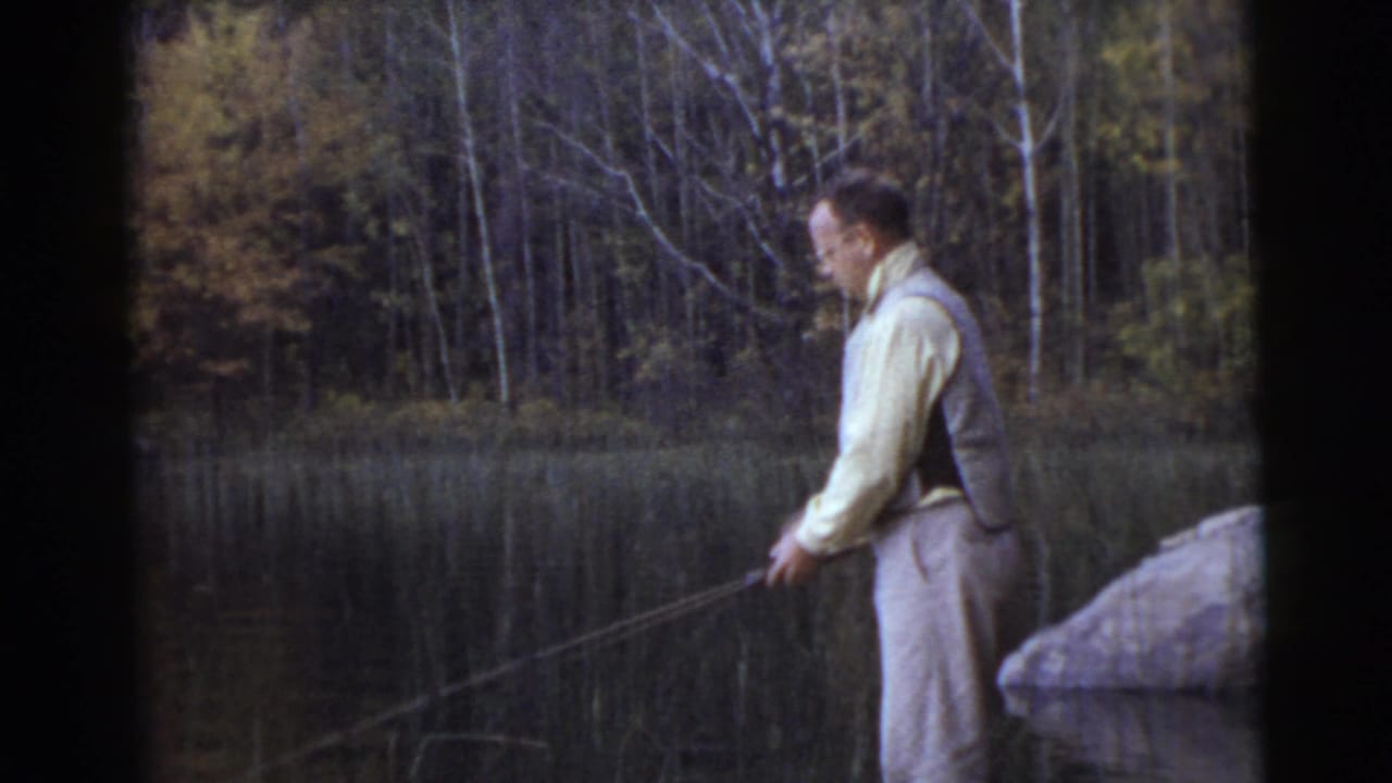 Man stands in profile with a fishing rod extended over lake water.