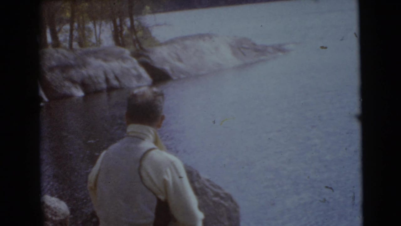 Man in a light vest stands near rocks and water with trees along the shore.