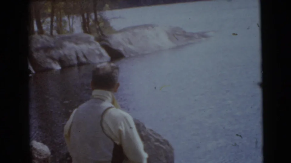1956 Miami home-movie frame showing a man from behind beside a rocky waterside with trees and calm water.