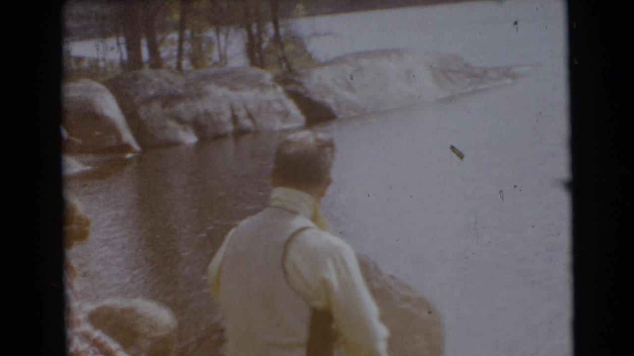 Man stands near the water as rocky shore and trees fill the background.