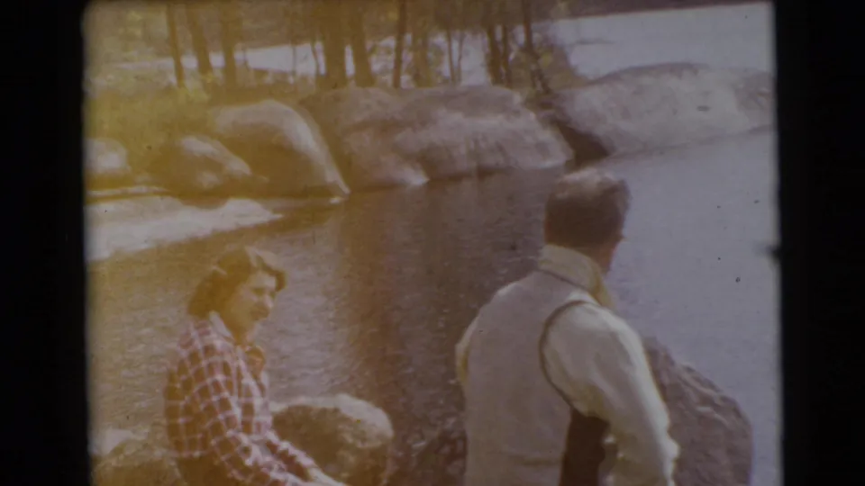 1956 Miami home-movie frame showing a seated woman in plaid and a standing man at a rocky waterside.