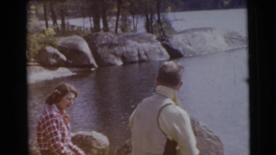 1956 Miami frame of a woman in plaid seated near boulders and a man standing by calm water.
