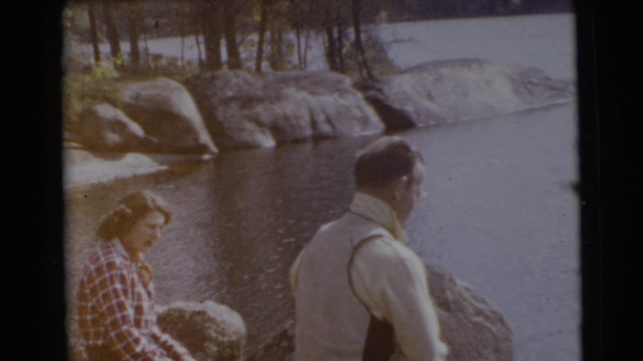 Two adults sit on shoreline rocks beside calm water and large boulders.