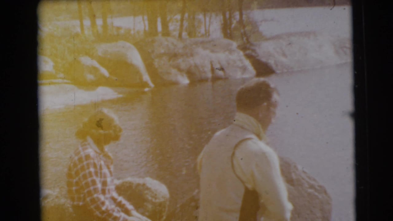 A yellow-tinted frame shows two adults seated near water and boulders.