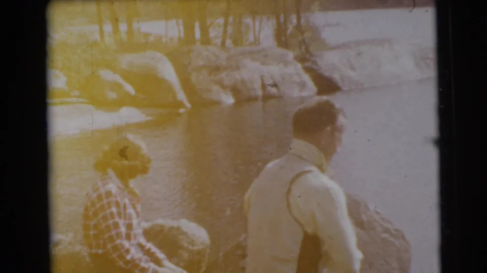 Warm, overexposed 8mm frame of two adults by a Miami lakeside, with rocks, trees, and water in view.