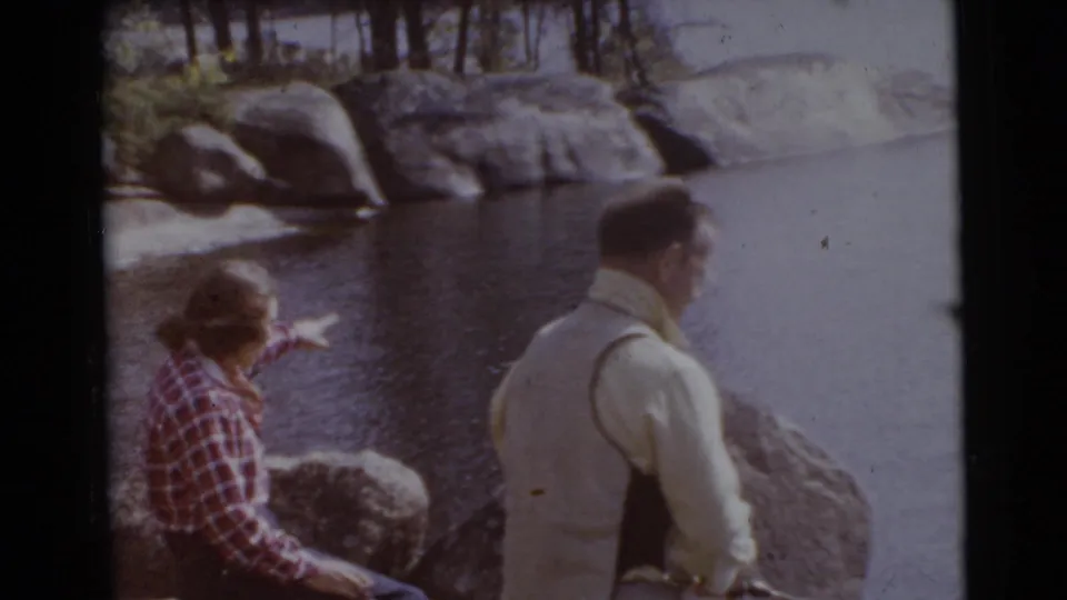 The woman in plaid points or gestures across the lakeside while the man in a yellow shirt looks toward the water.