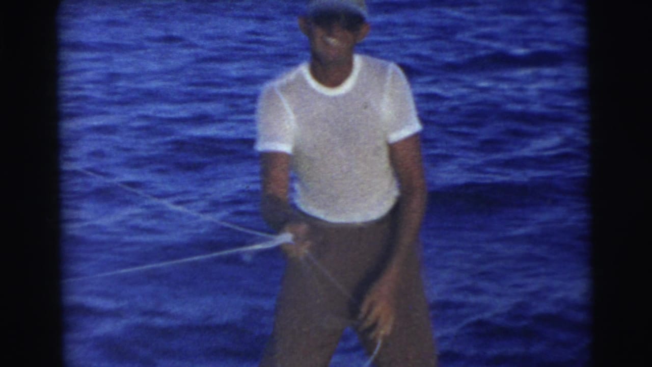 Man in a white shirt and cap stands on a boat holding a thin rope over blue water.
