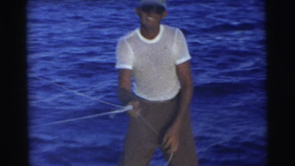 In 1956 Miami footage, a man in a white shirt and cap stands aboard a boat while handling a thin rope over open water.