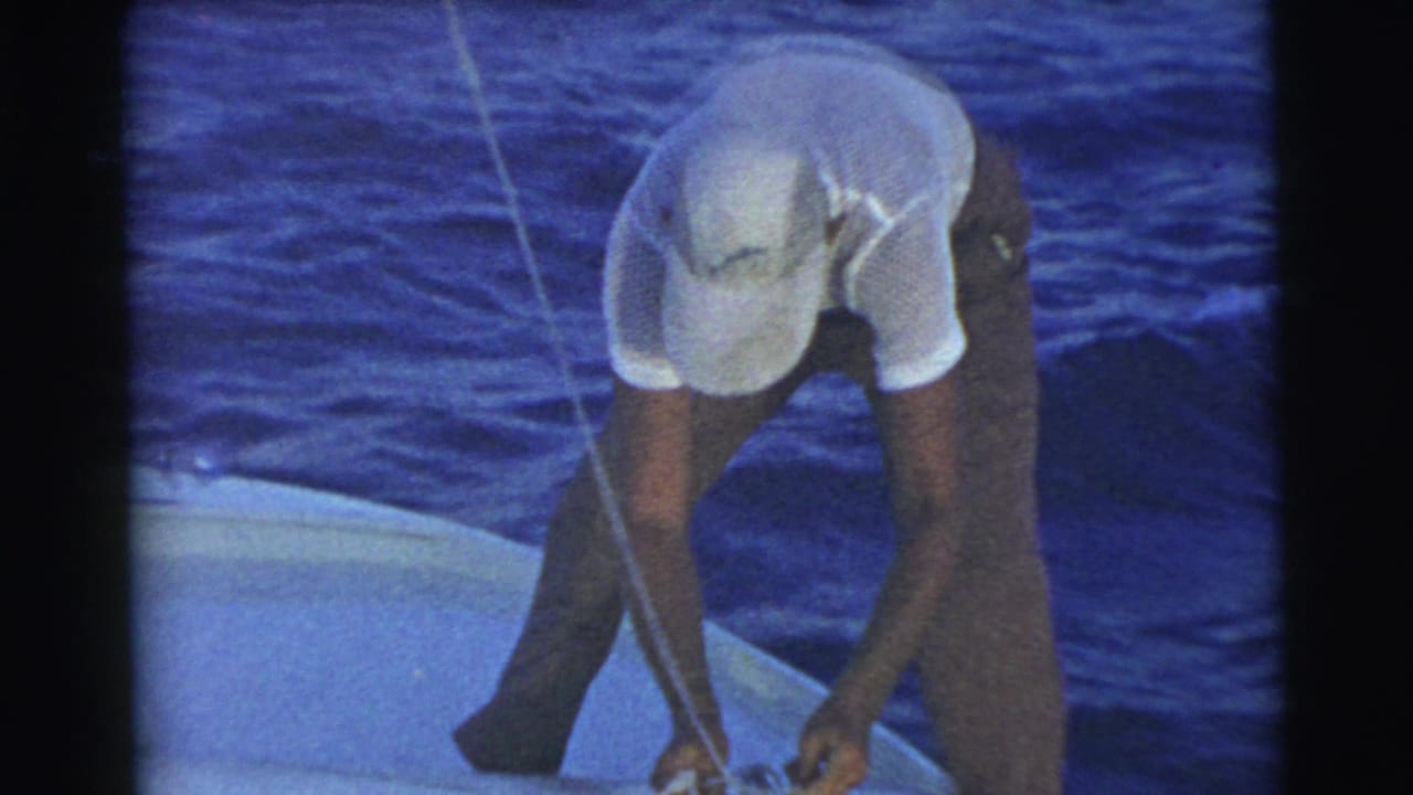 Man in a white cap bends over rope on a boat beside dark blue water.