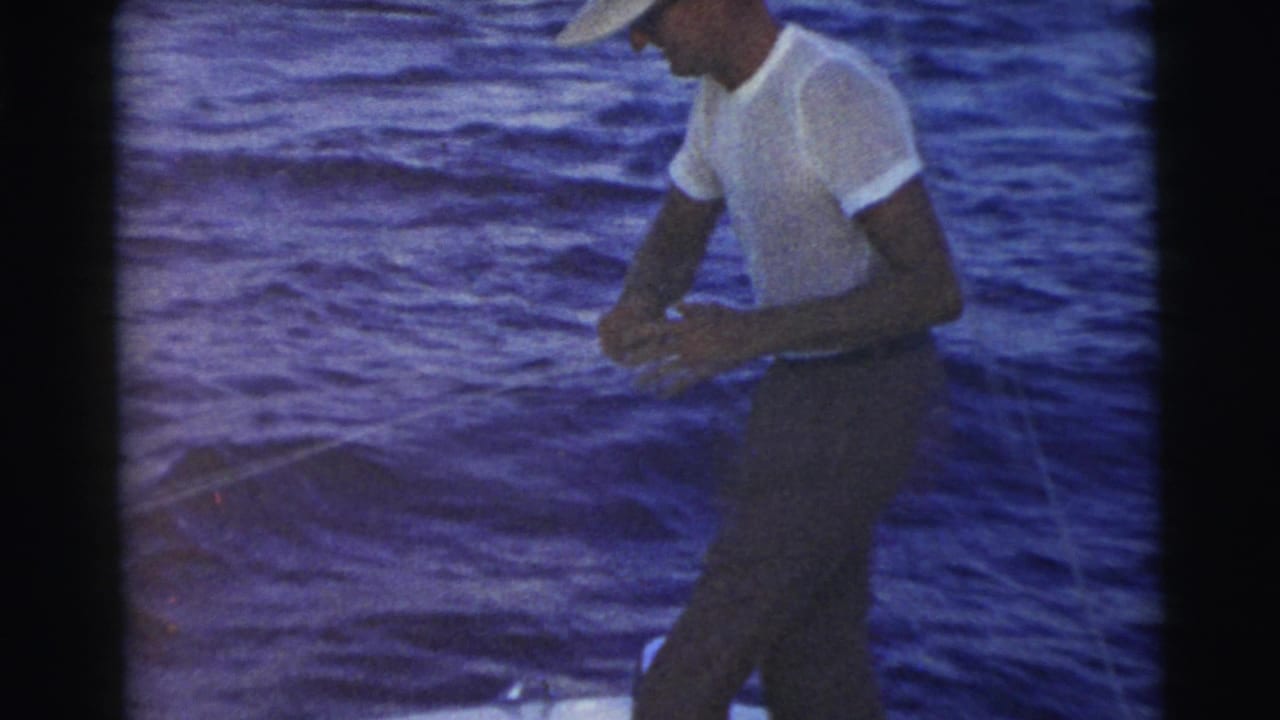 A man in a white cap and T-shirt stands on a boat against blue water in the Miami 1956 home-movie clip.