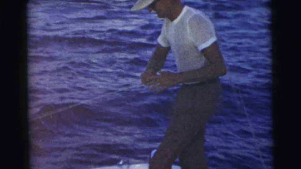 A man in a white cap and T-shirt stands on a boat against blue water in the Miami 1956 home-movie clip.
