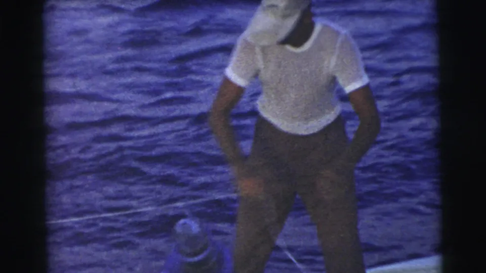 The man looks down as he handles a line near a fitting on the boat, with blue water behind him.