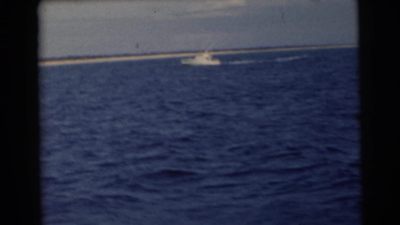 Blue sea with a narrow sandy island on the horizon and a small white motorboat near shore.