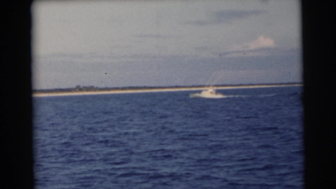 White motorboat crossing blue water in front of a low sandy island under a pale sky.