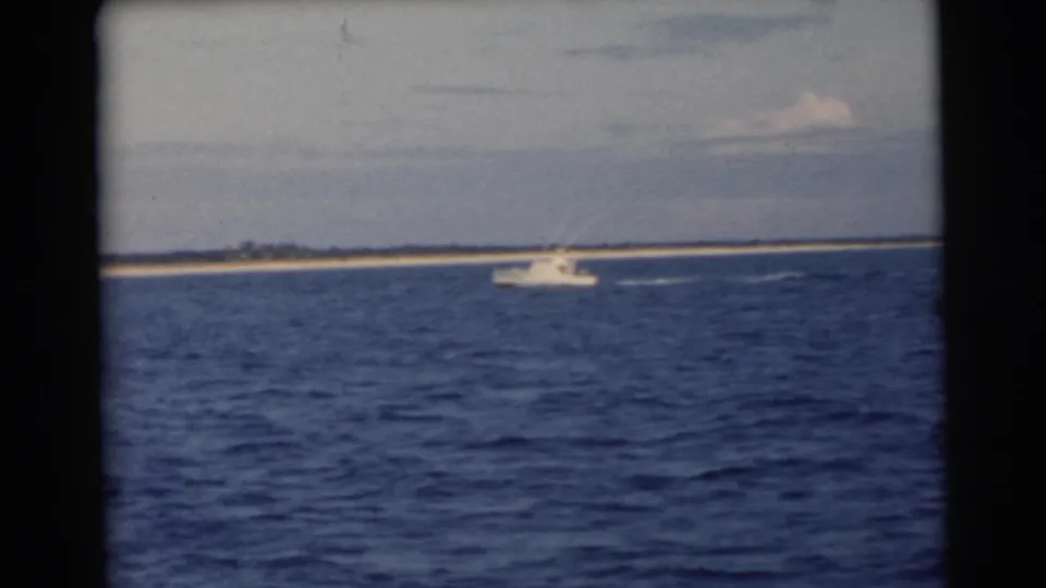 1956 Miami-area 8mm frame showing a white motorboat passing before a narrow island, with wake trailing across the blue sea.