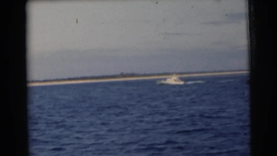 1956 Miami-area home-movie frame with rippled blue water, a distant white motorboat, and a low sandy island on the horizon.