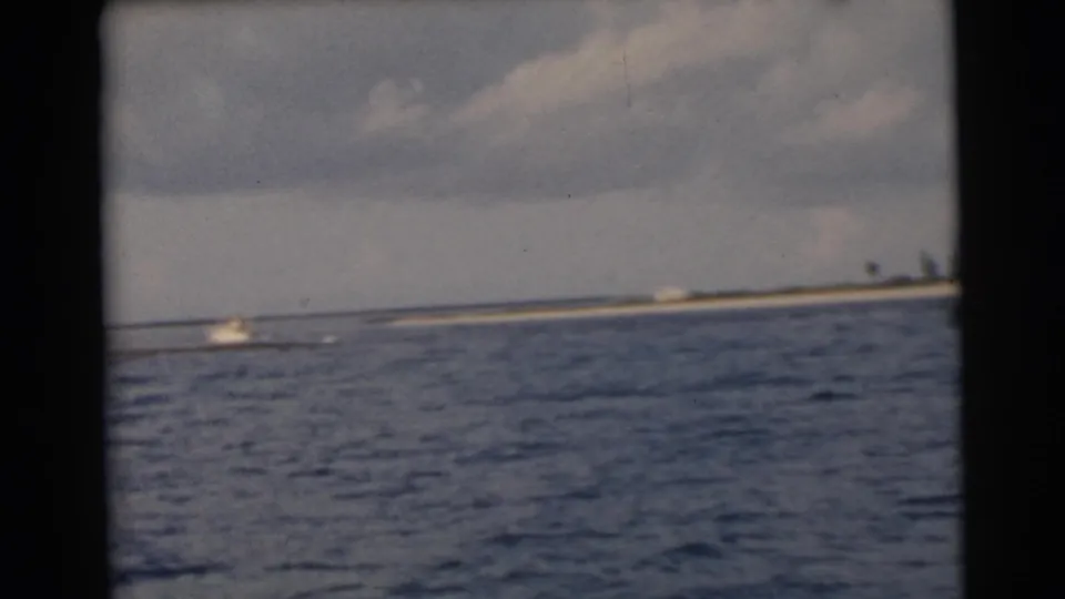 A small boat travels across choppy water near the Miami coast, filmed from a moving 8mm home-movie camera.