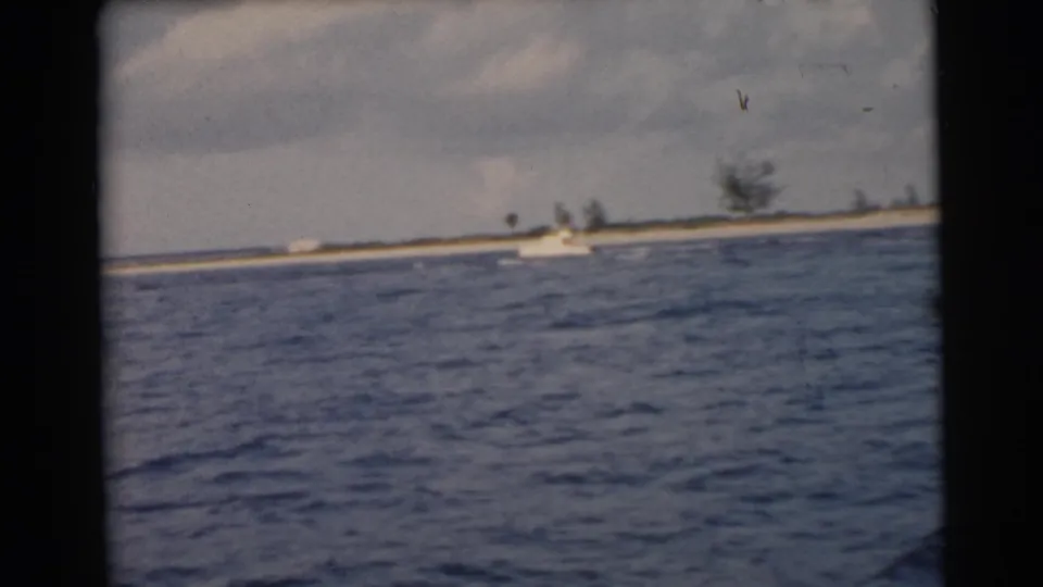 1956 Miami-area 8mm frame showing blue water from a moving boat, with a low sandy shoreline and sparse vegetation in the distance.