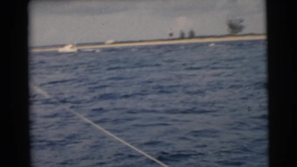 1956 Miami-area boating frame with blue water, a diagonal wake line, and a low sandy shoreline with sparse trees in the distance.