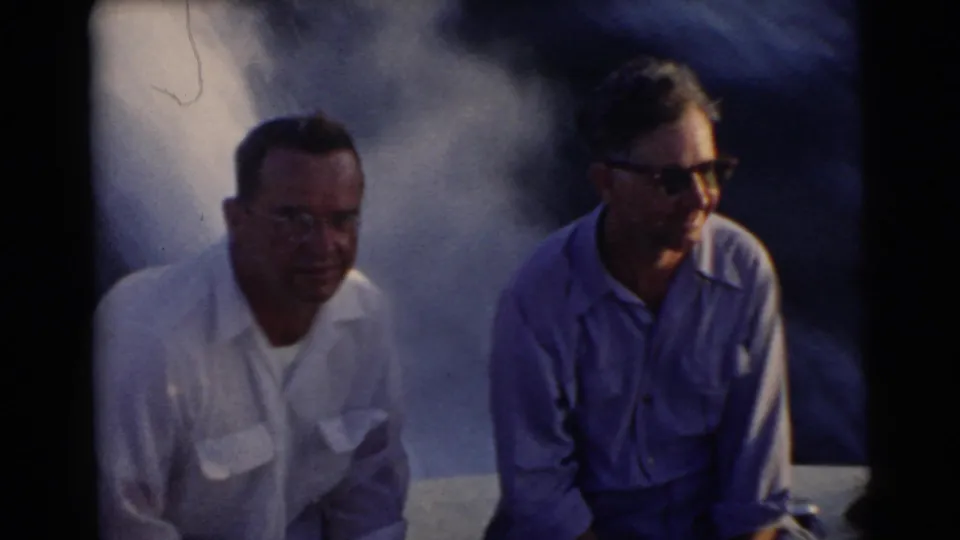 Two adult men in casual shirts ride in a boat in 1956 Miami-area footage, with water spray or wake behind them.