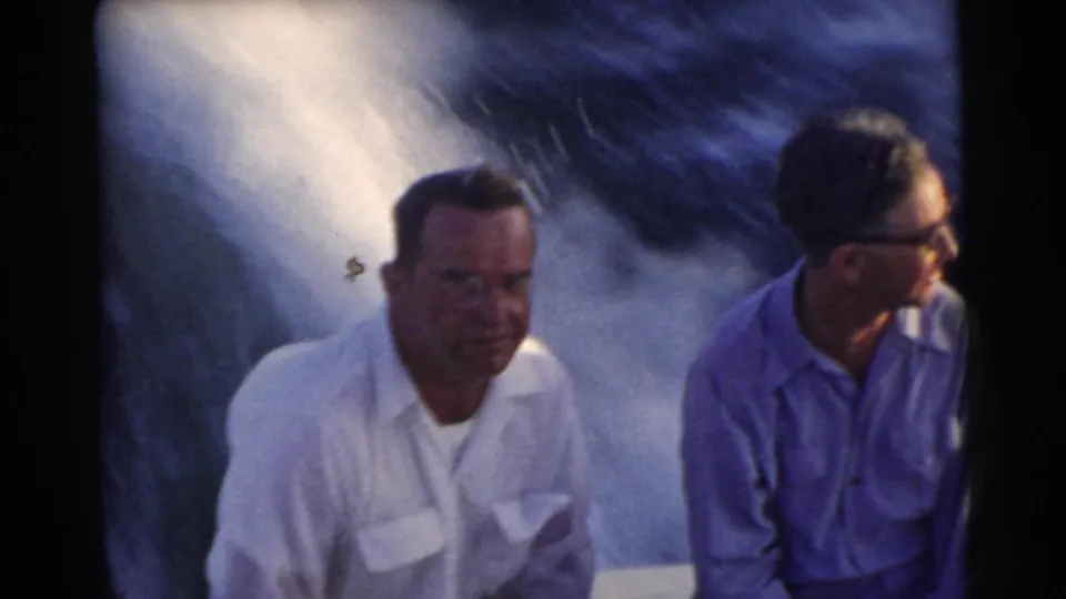 1956 Miami-area 8mm frame of two adult men riding in a moving boat, one facing camera and one looking aside.