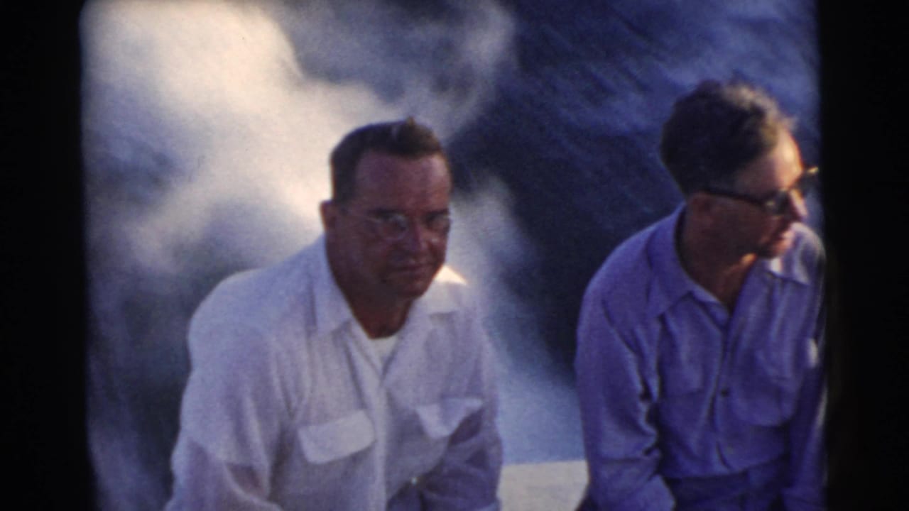 Two adult men in glasses sit at the back of a moving boat with water spray behind them.