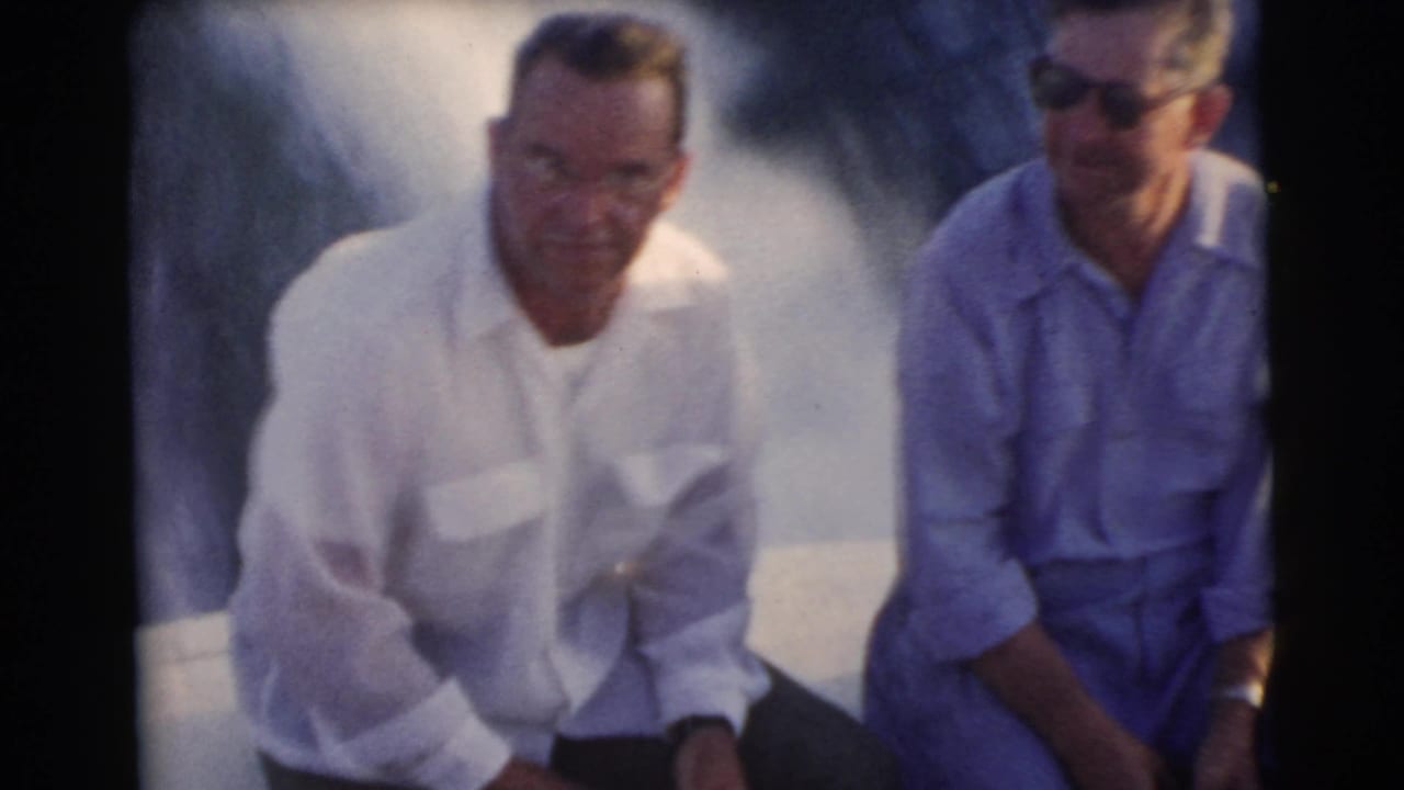 Two adult men wearing glasses sit on the back of a moving boat in Miami, with bright spray and soft 8mm color visible around them.