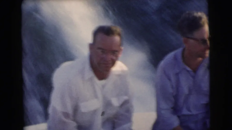 A close, soft 8mm frame shows two adult men seated on a moving boat in 1956 Miami, with spray behind them and the right figure partly cropped.