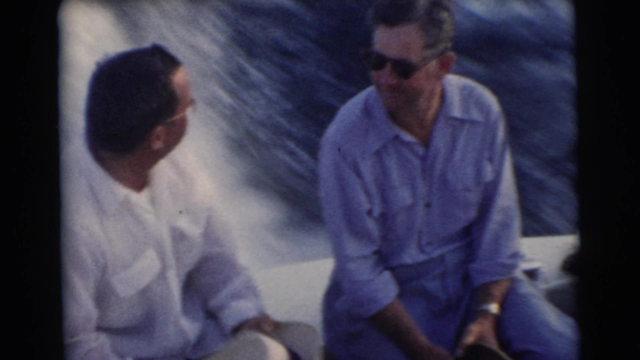 Two adult men sit on a boat and talk, with water behind them.