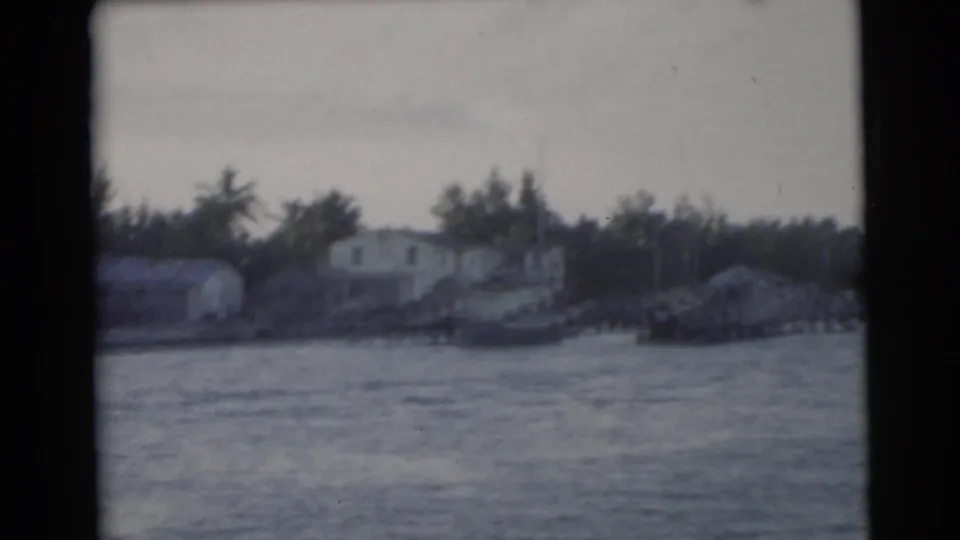 1956 Miami home-movie frame with low waterfront houses, small boats or docks, rippled water, and dark trees along the shore.