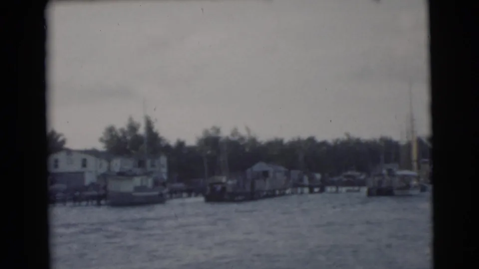 1956 Miami shoreline frame showing moored boats, dock structures, low waterfront buildings, trees, and rippled water from a boat perspective.