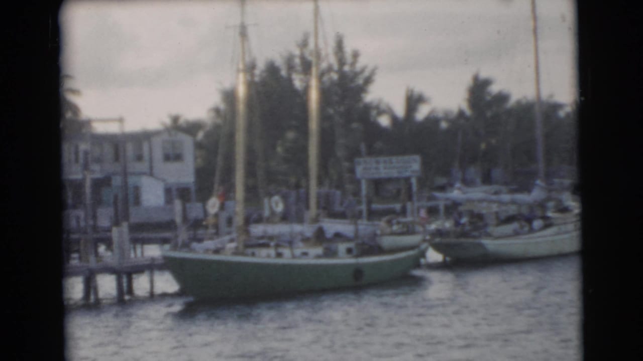 1956 Miami waterfront view with a green-hulled sailboat, neighboring boat, dock pilings, palms, and small shore structures.