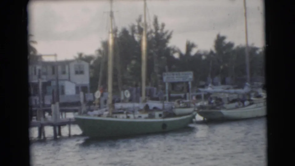 1956 Miami waterfront view with a green-hulled sailboat, neighboring boat, dock pilings, palms, and small shore structures.