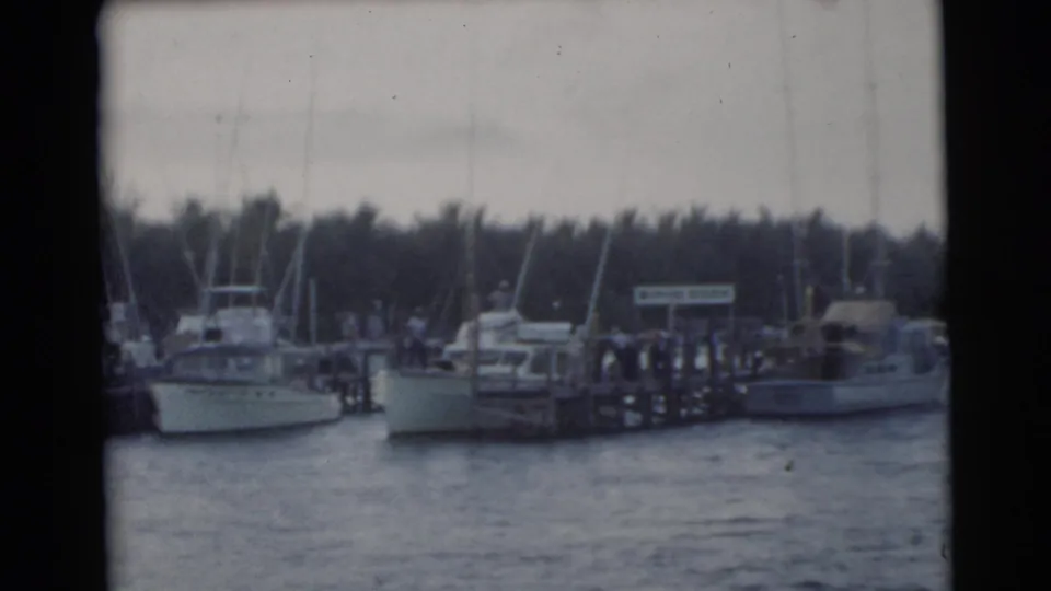 1956 Miami waterfront frame showing boats at a marina dock, with small indistinct figures near the pier and palm trees beyond.