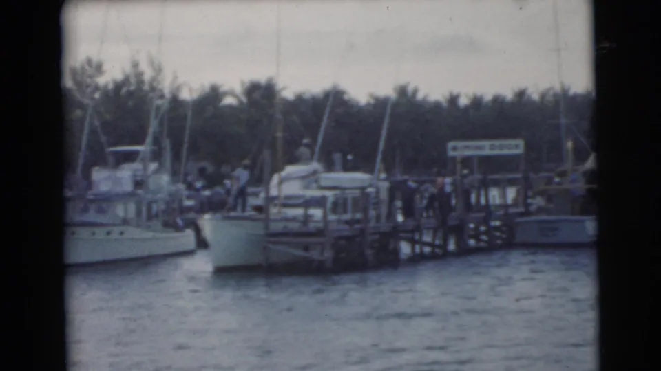 1956 Miami home-movie frame of a marina dock with moored boats, a sign on the pier, small figures, and palm trees in back.