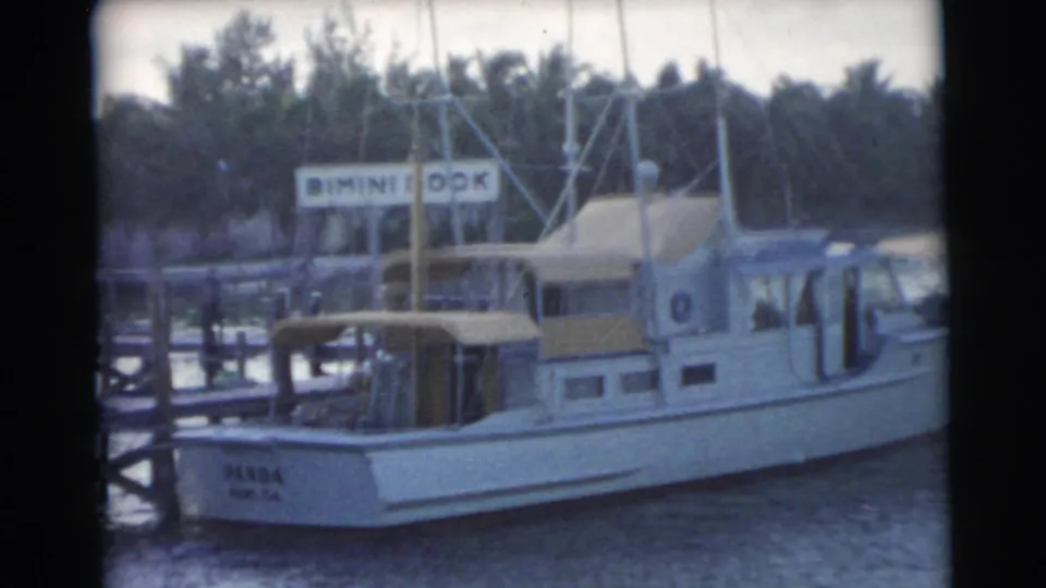 1956 Miami marina frame showing a light-colored boat moored at a wooden pier below the Bimini Dock sign and palm-lined background.