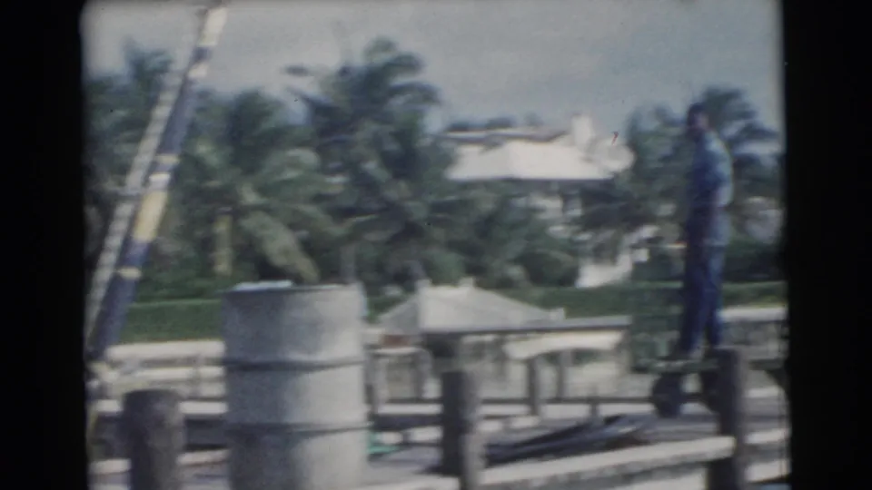 A 1956 Miami home-movie frame shows the man and green dolly along the dock, with palm trees, railings, and waterfront buildings beyond.