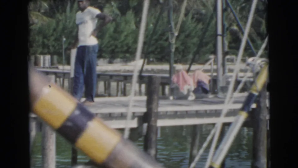 1956 Miami home-movie frame of a man standing on a wooden dock over water with palms and fishing poles around him.