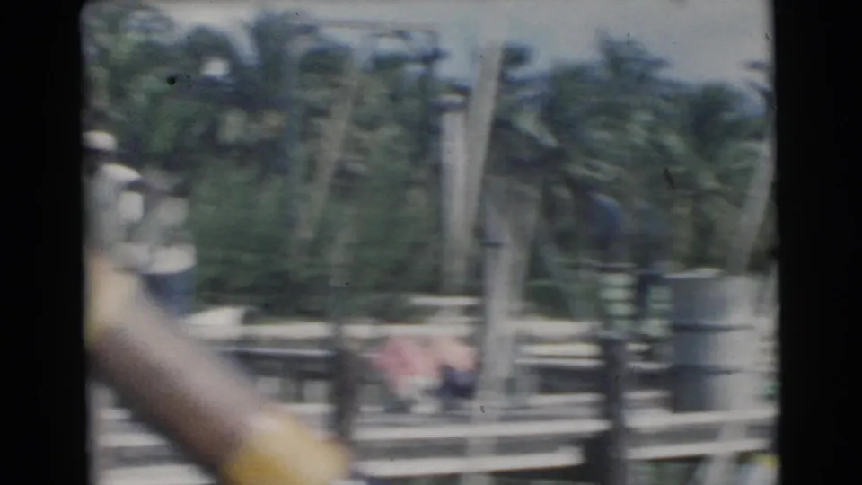 Soft 8mm frame from the Miami dock scene, with palms, railings, a barrel and a partially visible adult figure near the edge.