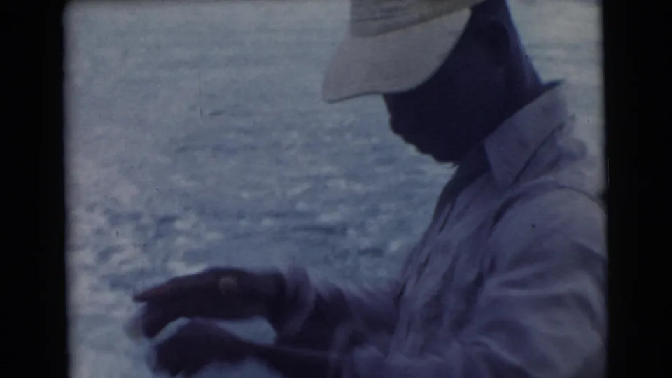 A man in a light cap and shirt stands near the water in profile, filmed from close range in the 1956 Miami-area clip.